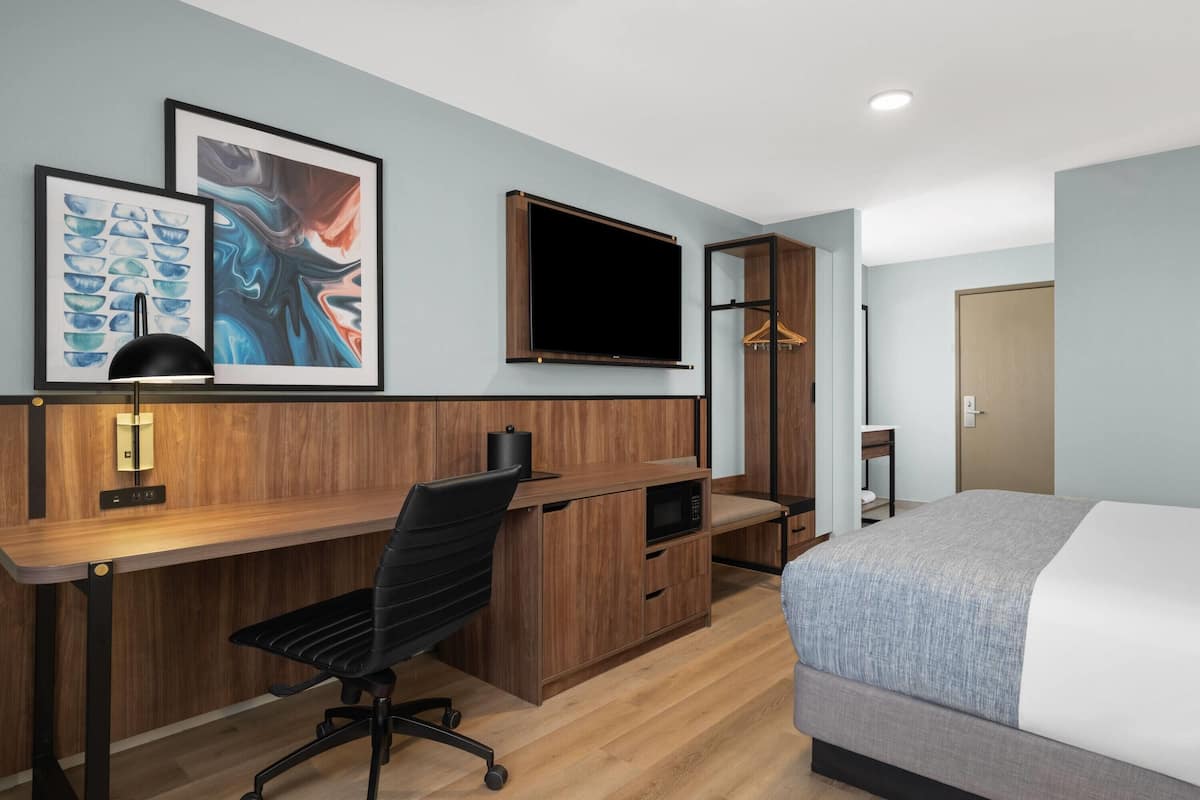 Quarto de hotel com escrivaninha de madeira, cadeira de escritório preta, cama com manta cinzenta, televisão de tela plana fixada na parede e duas obras de arte emolduradas sobre a escrivaninha.