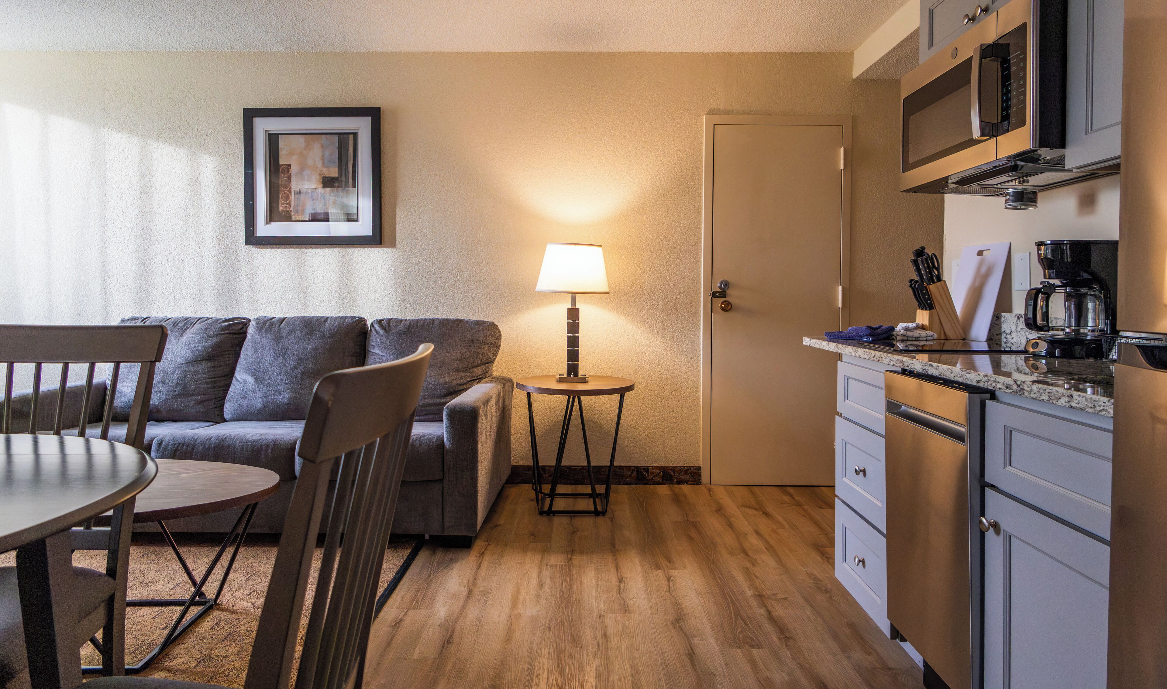 A compact living space with a dining table, chairs, a sofa, a small side table with a lamp, a kitchenette, and a framed picture on the wall.