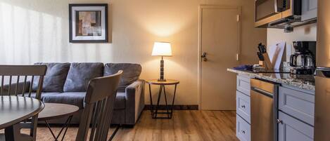 A compact living space with a dining table, chairs, a sofa, a small side table with a lamp, a kitchenette, and a framed picture on the wall.