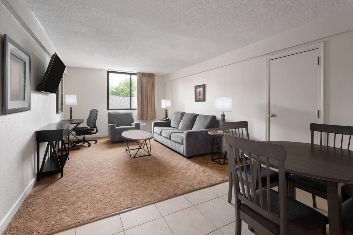 A modern hotel room with a sofa, a dining table, a TV mounted on the wall, and a large window.