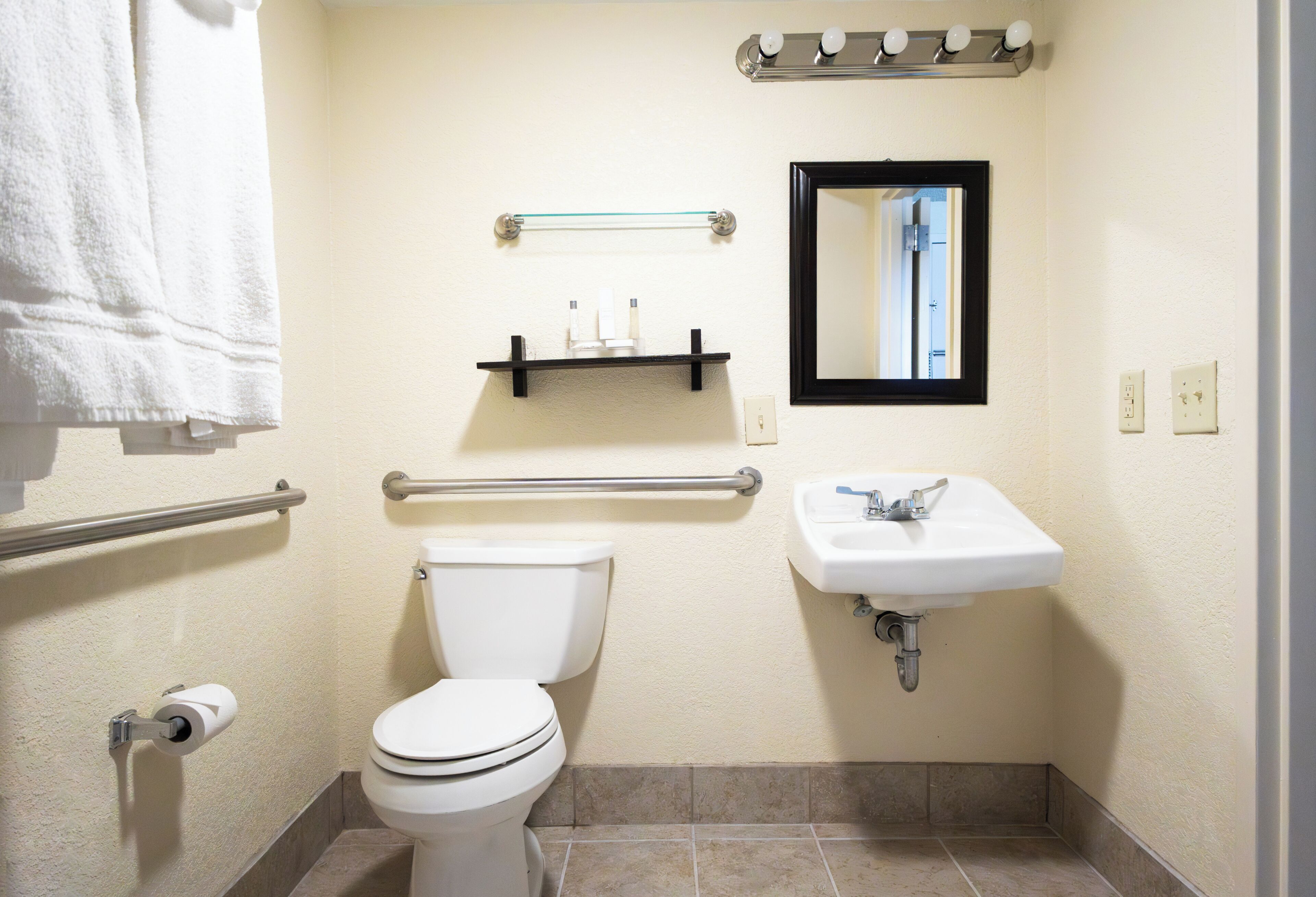 A standard bathroom with a toilet, sink, mirror, towel rack, and handrails.