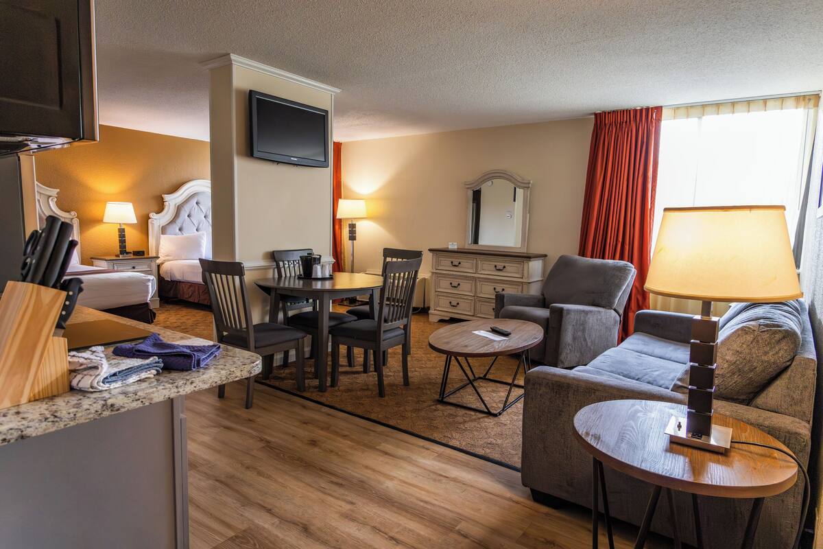 A hotel room with a bed, a dining table with chairs, a sofa, a TV, and a lamp.
