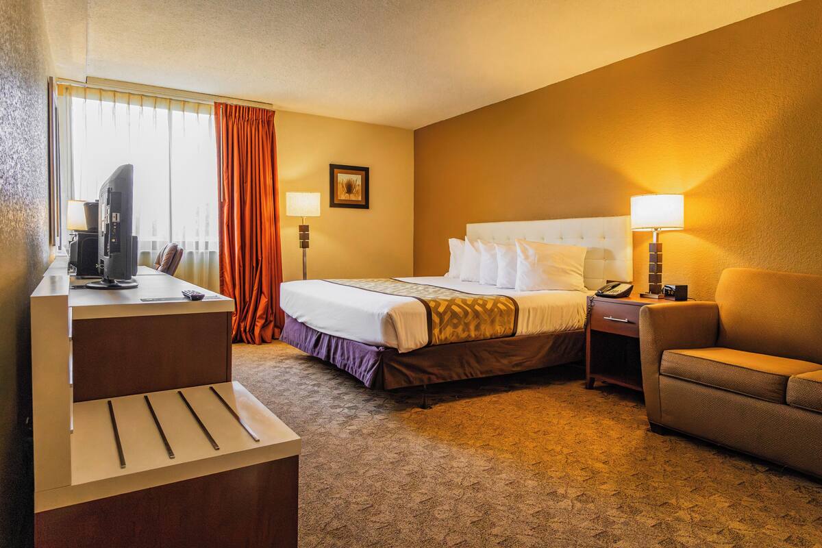 A hotel room with a bed, a desk with a television, a chair, and a nightstand with a lamp.