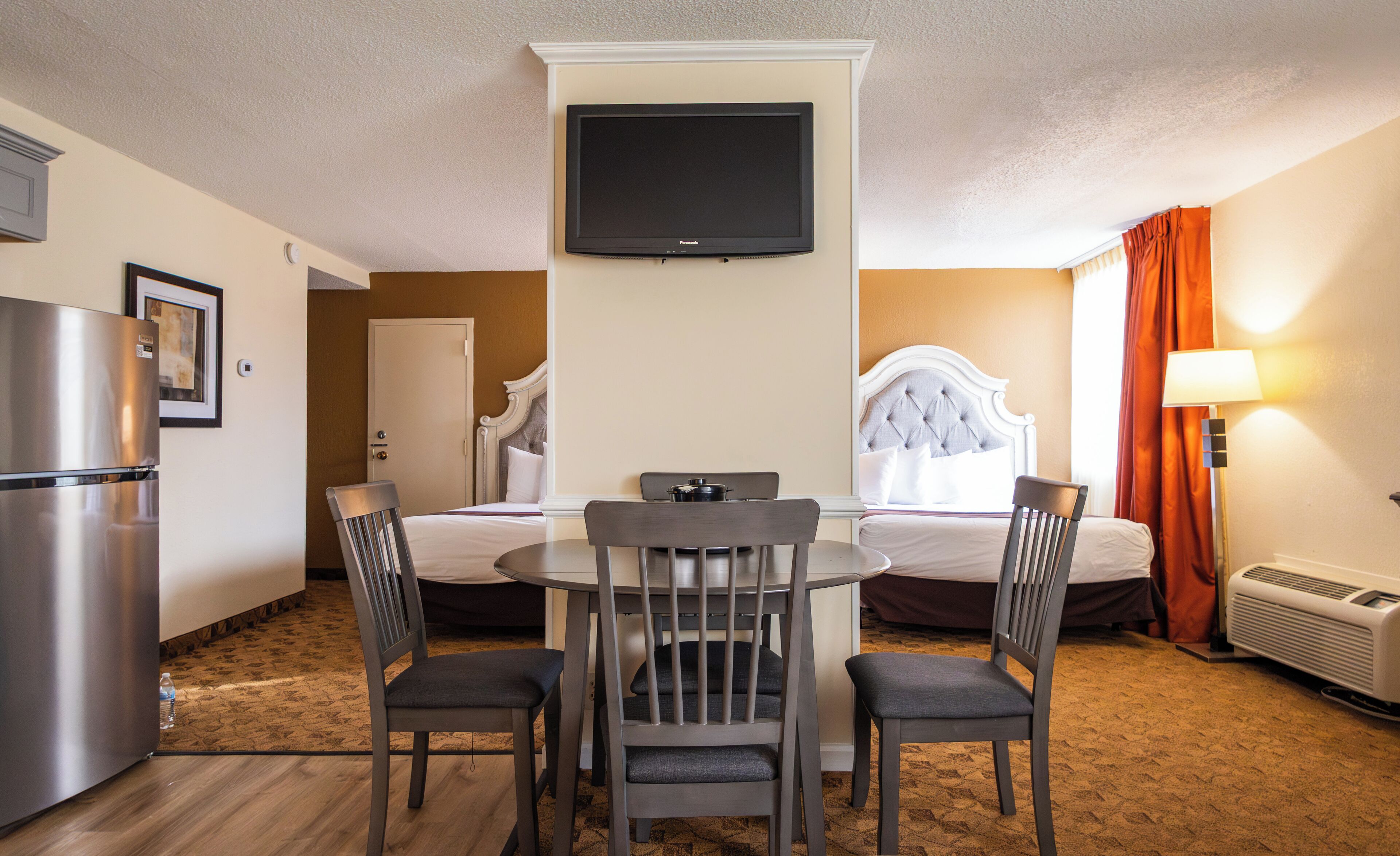 Una habitación de hotel con cama, televisión montada en la pared, mesa de comedor con sillas, refrigerador y aire acondicionado.