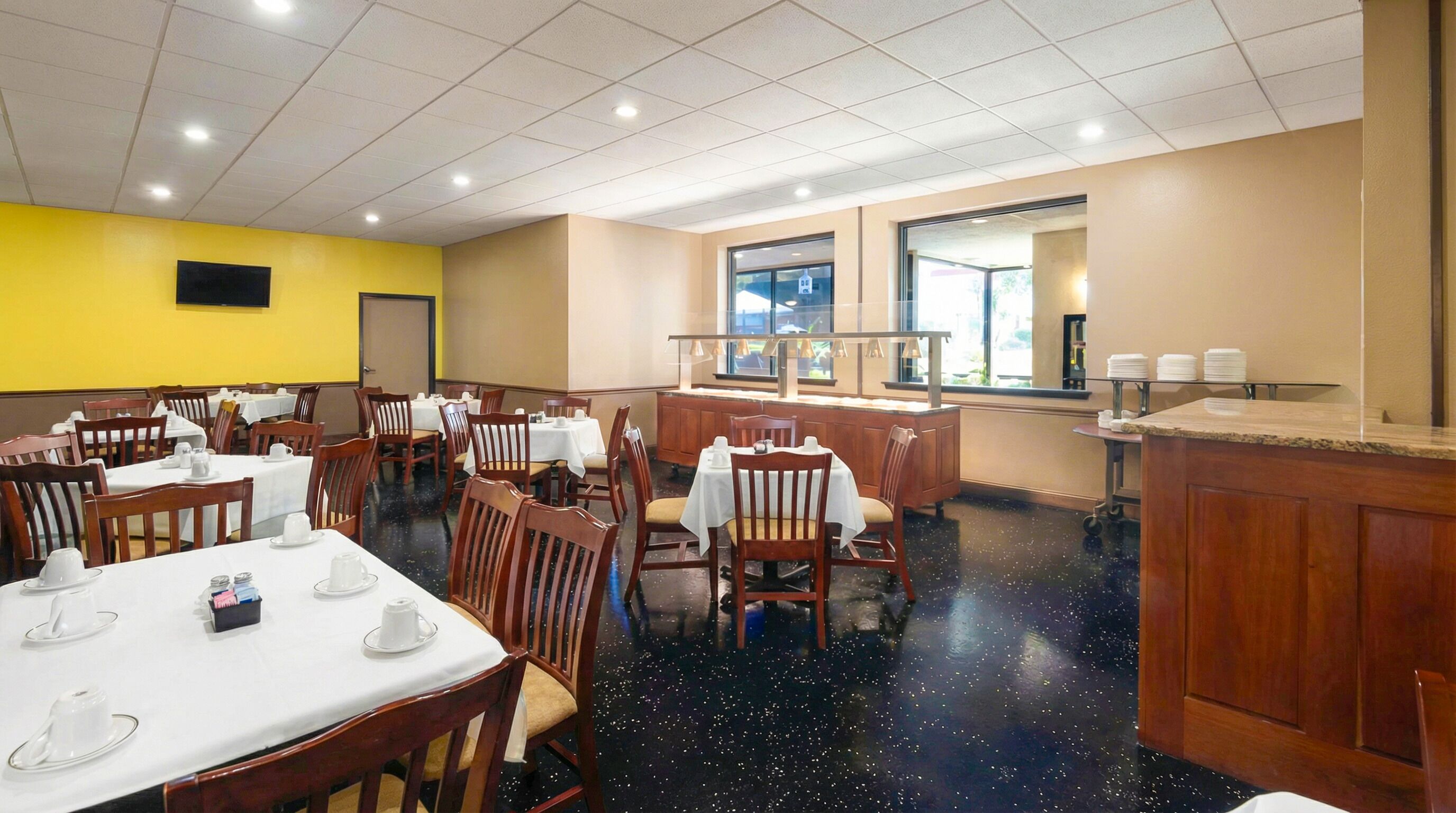 A dining area with tables set for service, a kitchen counter, and a large window providing a view.