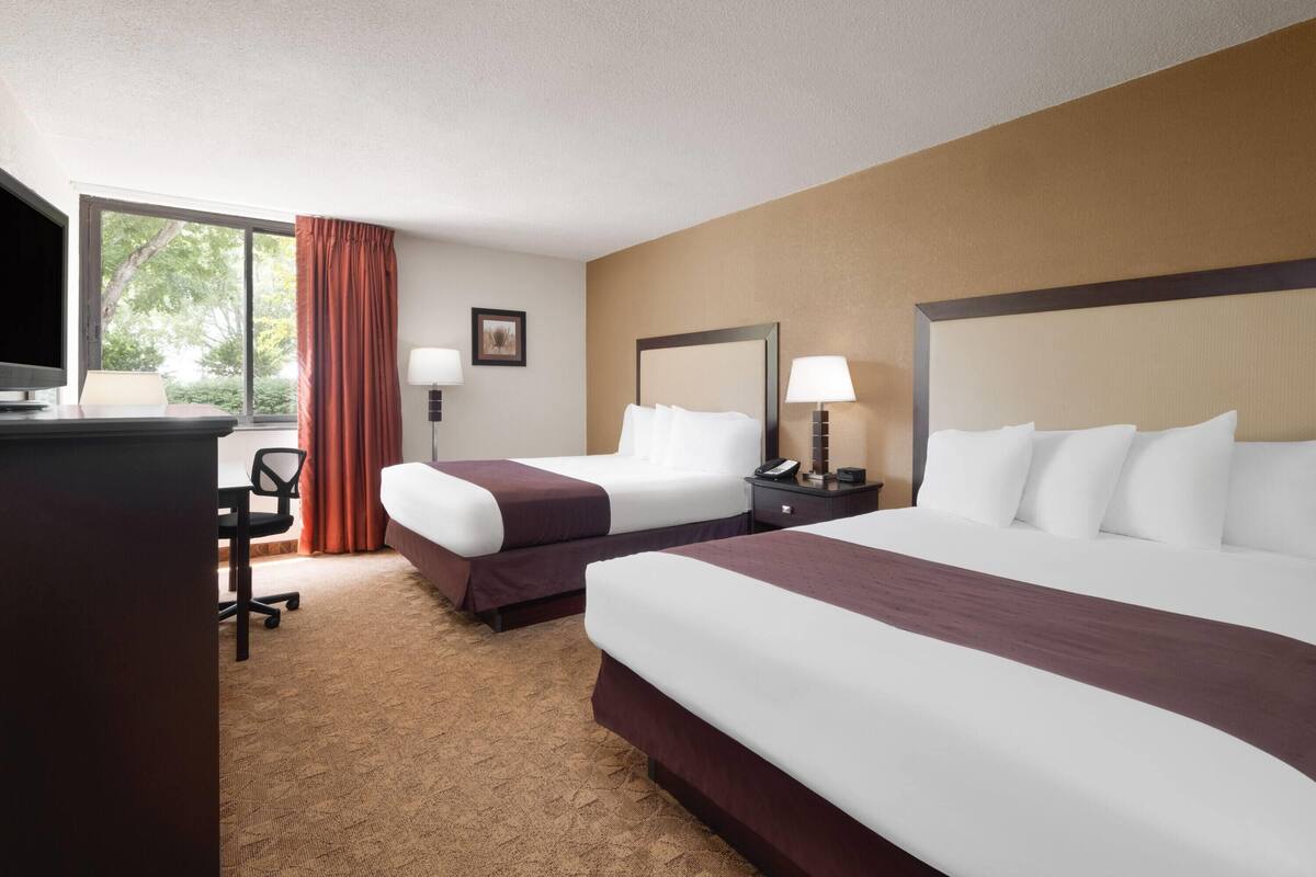 A hotel room with two beds, a desk, a chair, a TV, and a window with curtains.