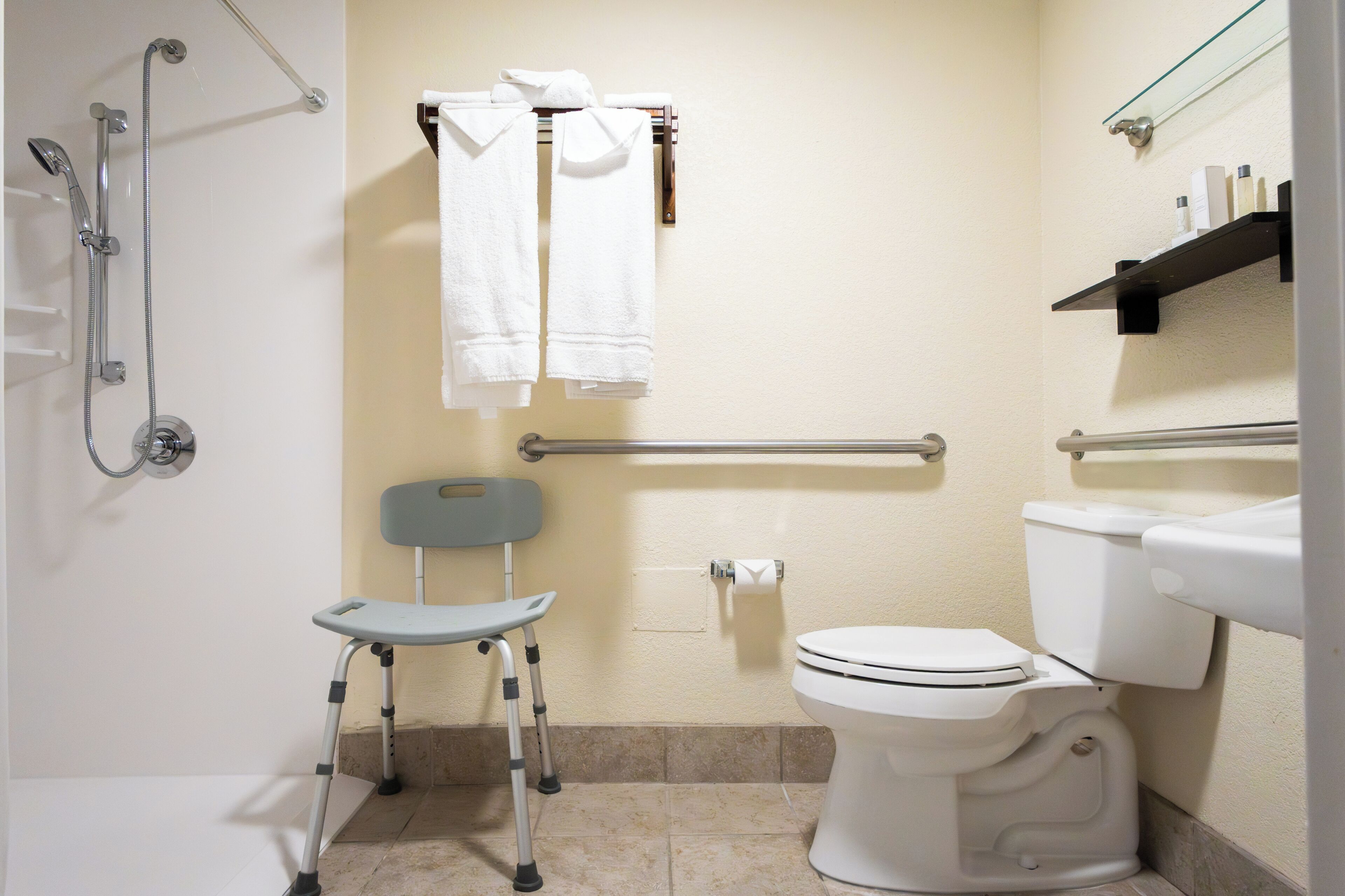 Un baño con ducha, inodoro, barra de apoyo, silla de ruedas y perchero para toallas.