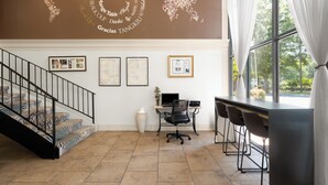 A hotel lobby with a staircase, a desk with chairs, framed pictures on the wall, and a large window with curtains. - Village Inn Clemmons/Winston Salem, Trademark by Wyndham (Clemmons)
