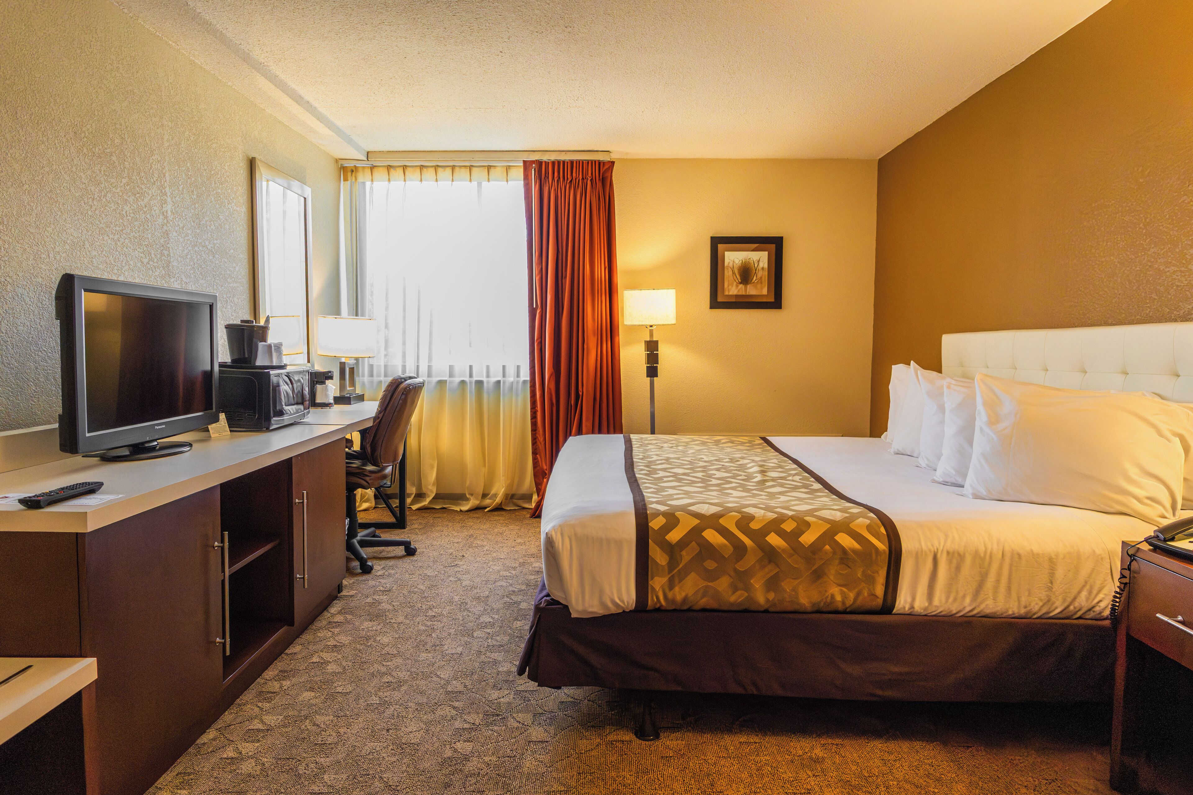 A hotel room with a bed, a desk with a television, a lamp, and a window with curtains.