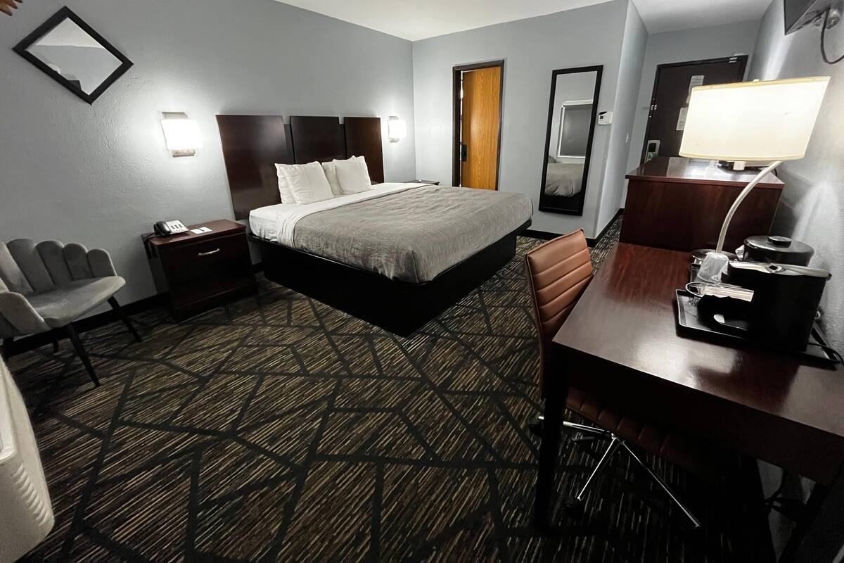 A hotel room with a bed, bedside table, chair, desk, lamp, and a mirror.