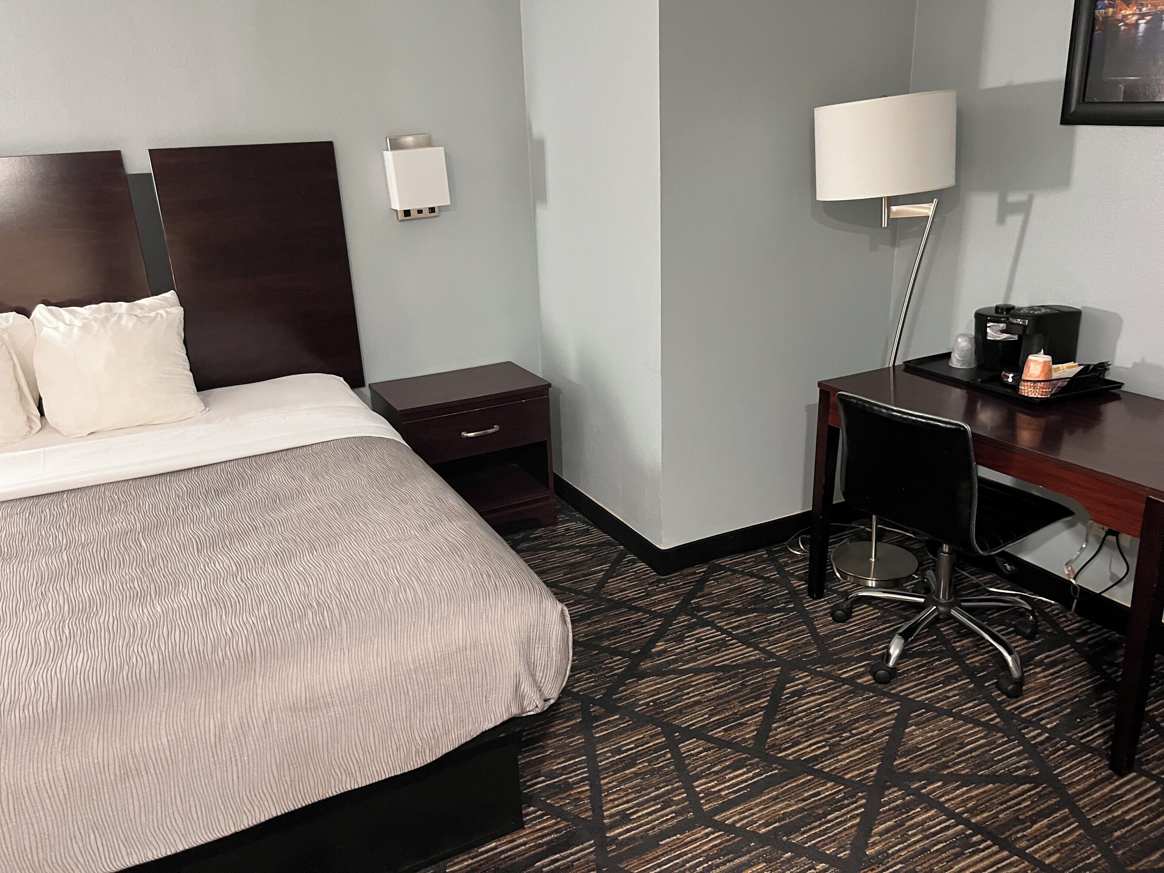 A hotel room with a bed, a desk, a chair, a nightstand, a lamp, and a coffee maker.