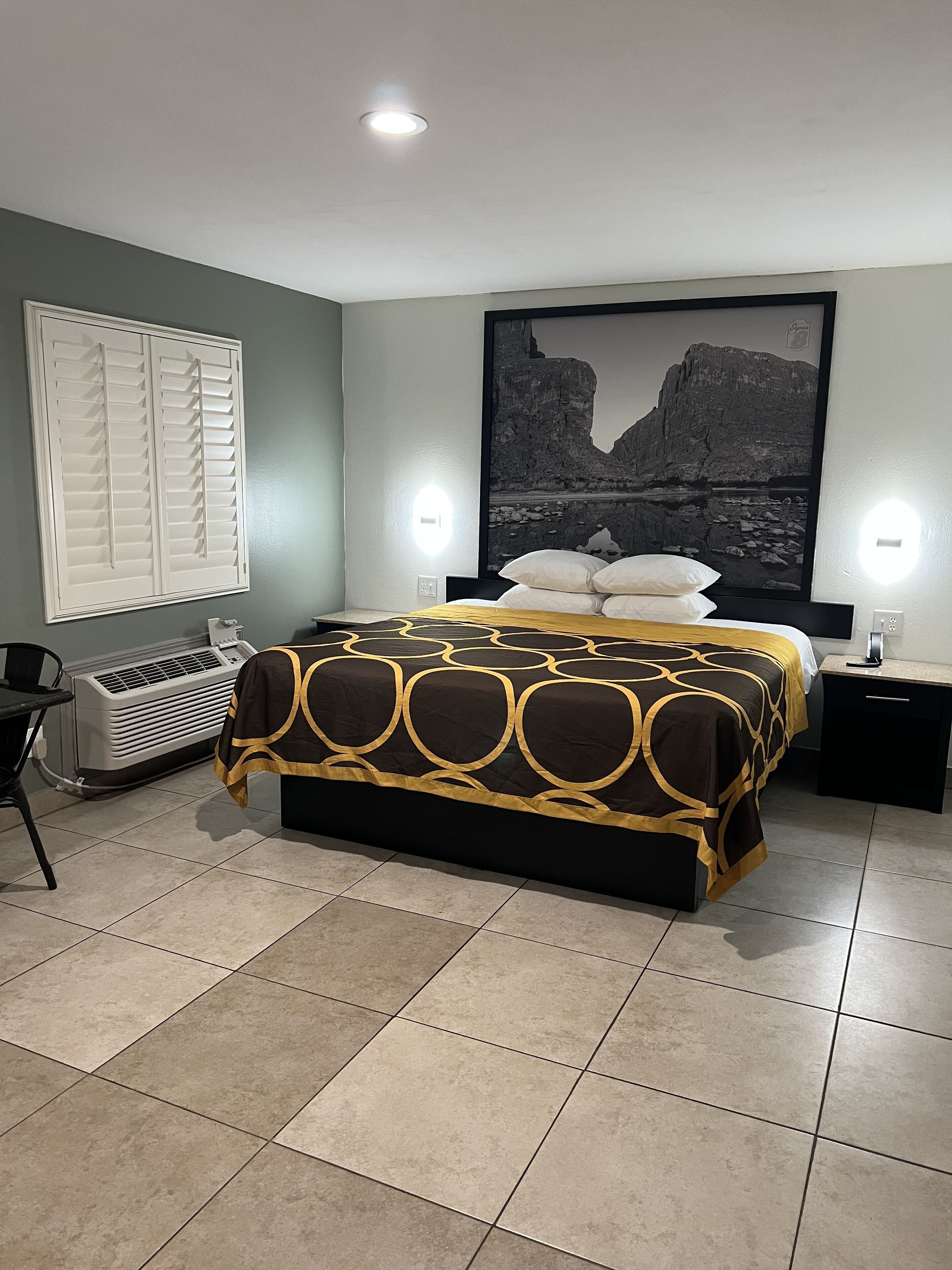 A bedroom with a bed, a window with blinds, a heater, a nightstand, and a wall-mounted light.