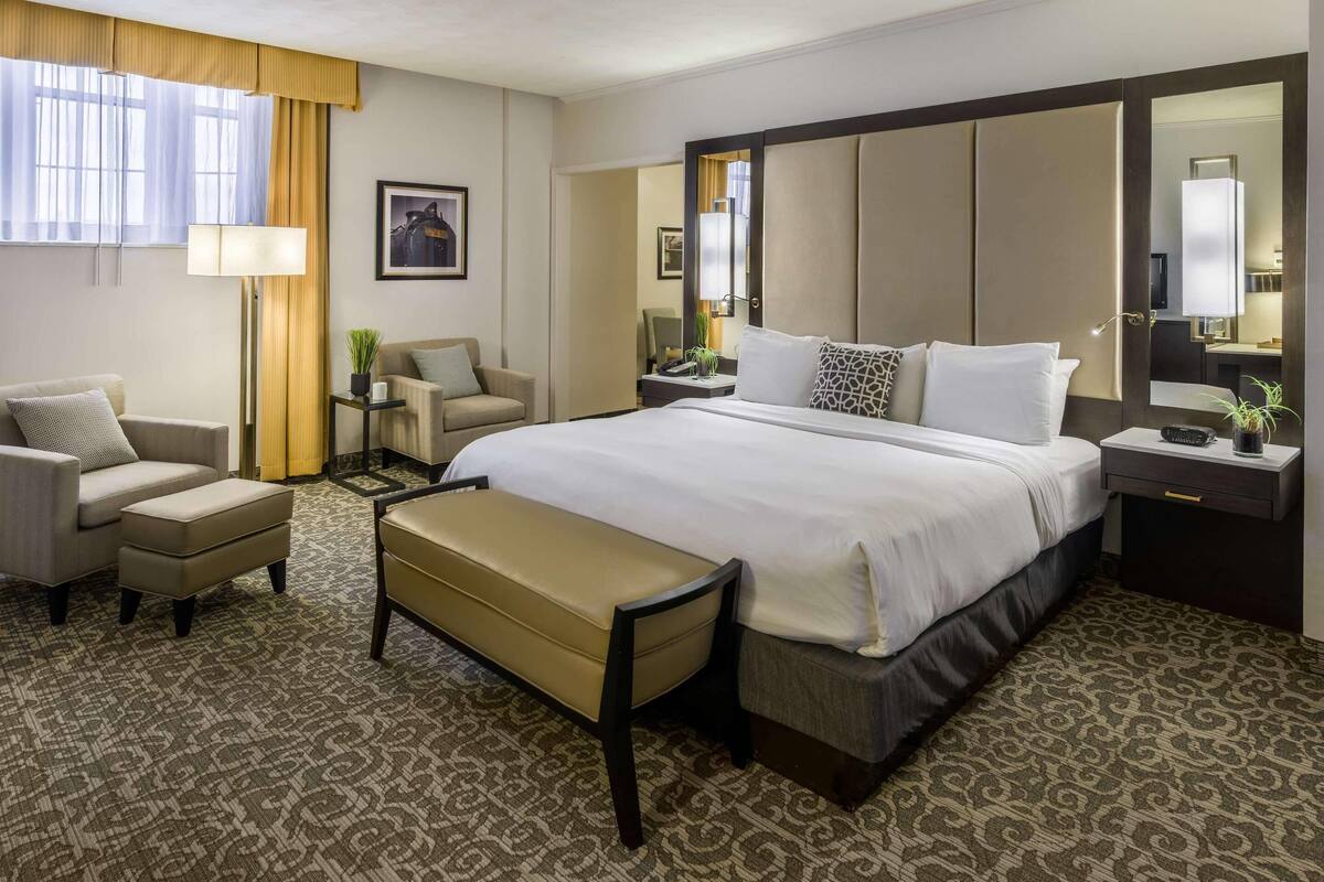 A hotel room with a large bed, a sofa, a chair, a desk with a lamp, and a mirror.