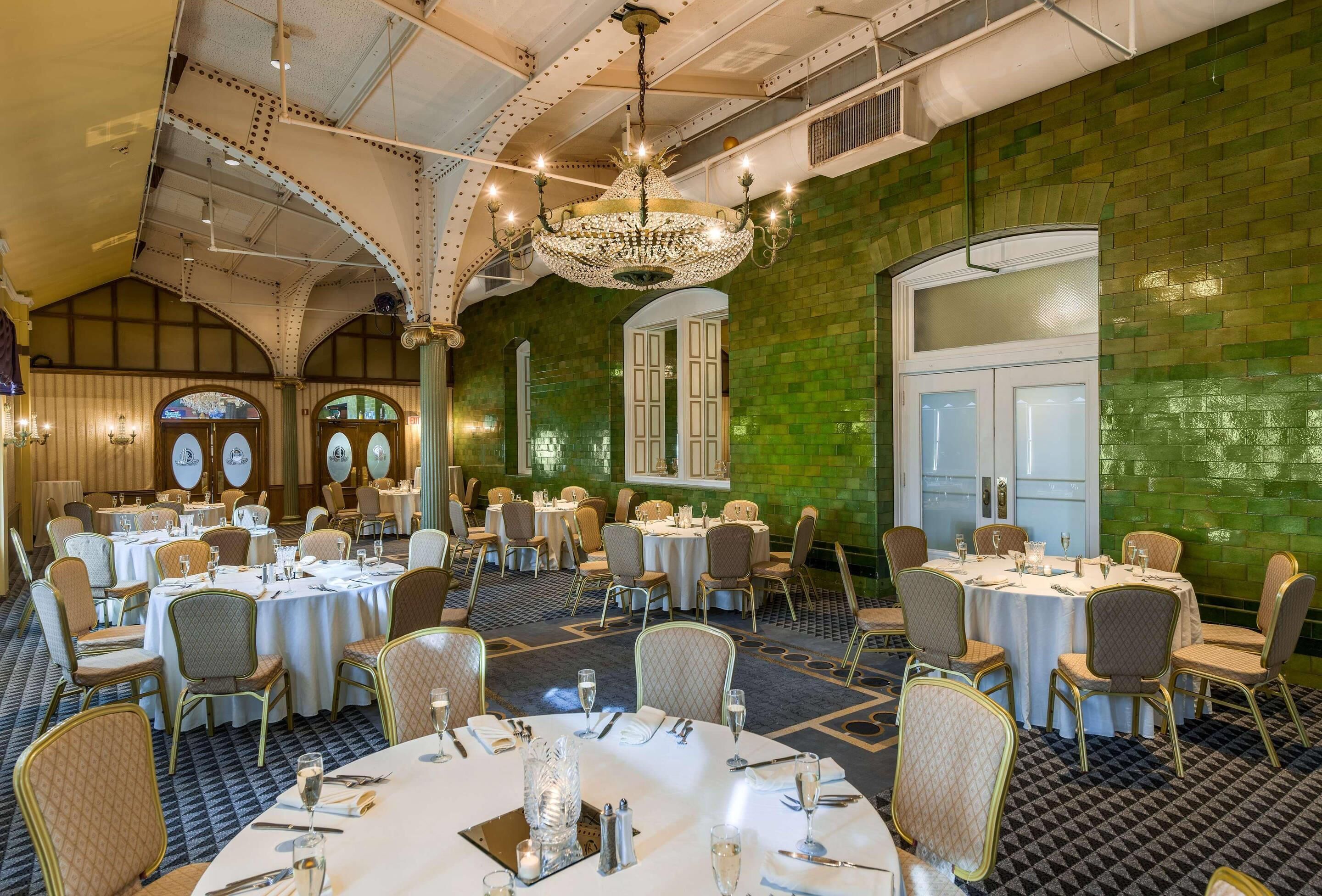 A banquet hall with round tables set for dining, a chandelier, and green-tiled walls.