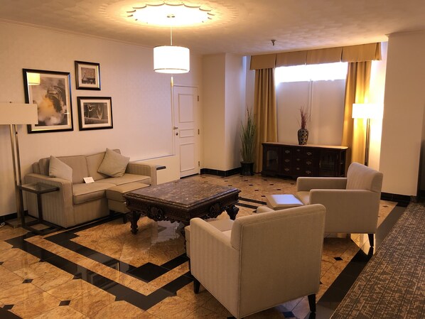 A hotel room with a sofa, armchairs, a coffee table, a lamp, a potted plant, and a view of the outside through a window with curtains.