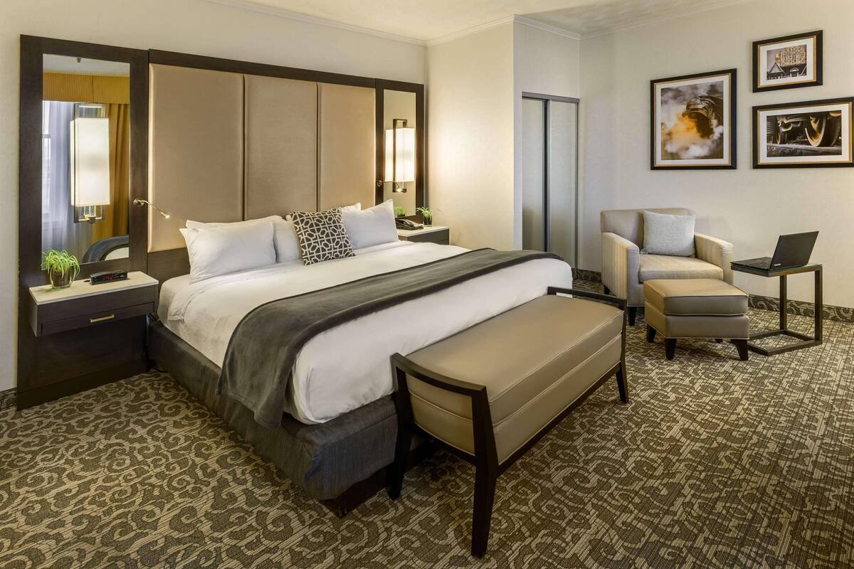 A hotel room with a large bed, a desk, a chair, a bench, and framed pictures on the wall.