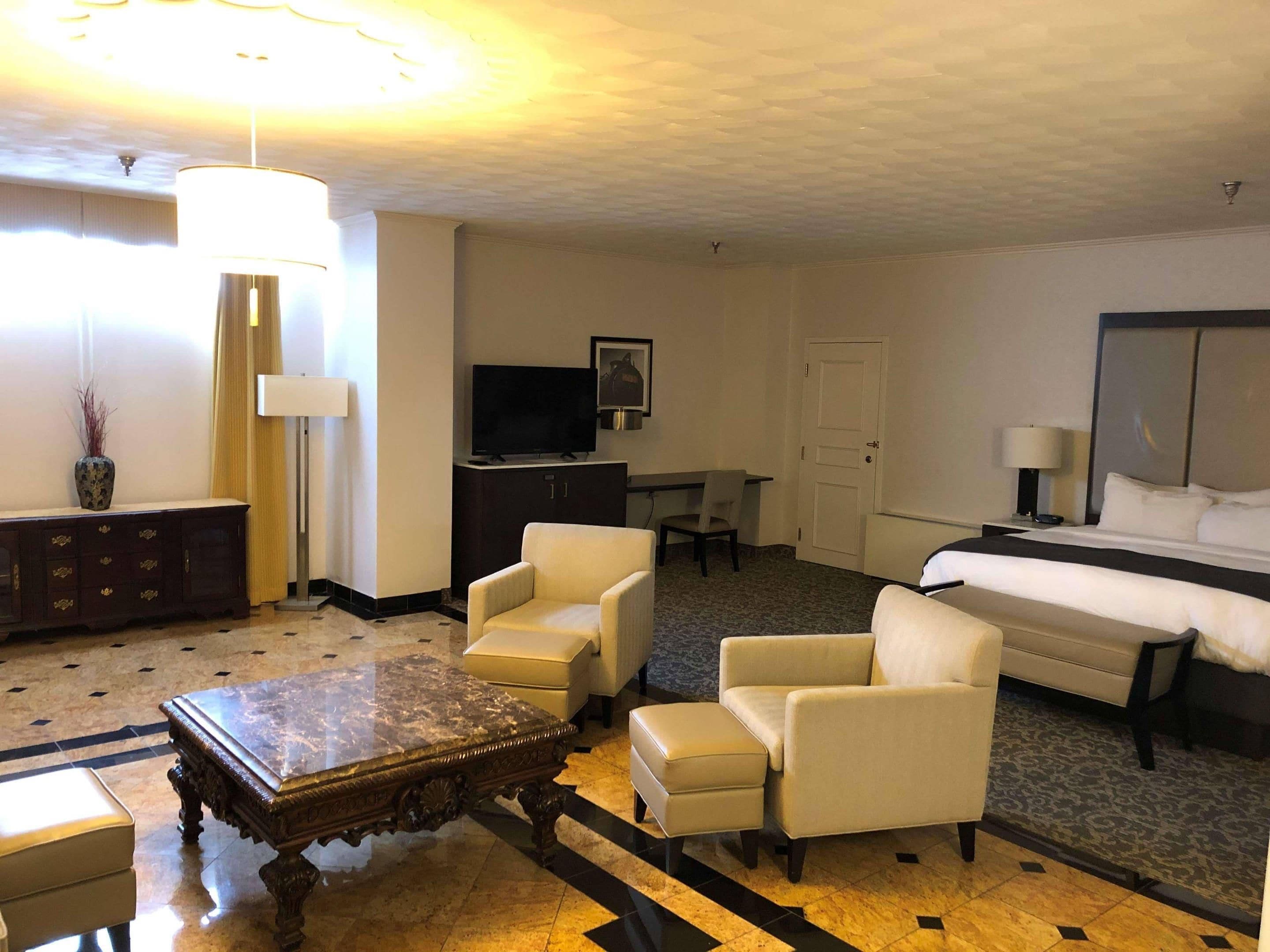A hotel room with a bed, two armchairs, a wooden coffee table, a TV, a desk, and a lamp.