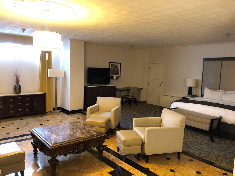 A hotel room with a bed, two armchairs, a wooden coffee table, a TV, a desk, and a lamp.