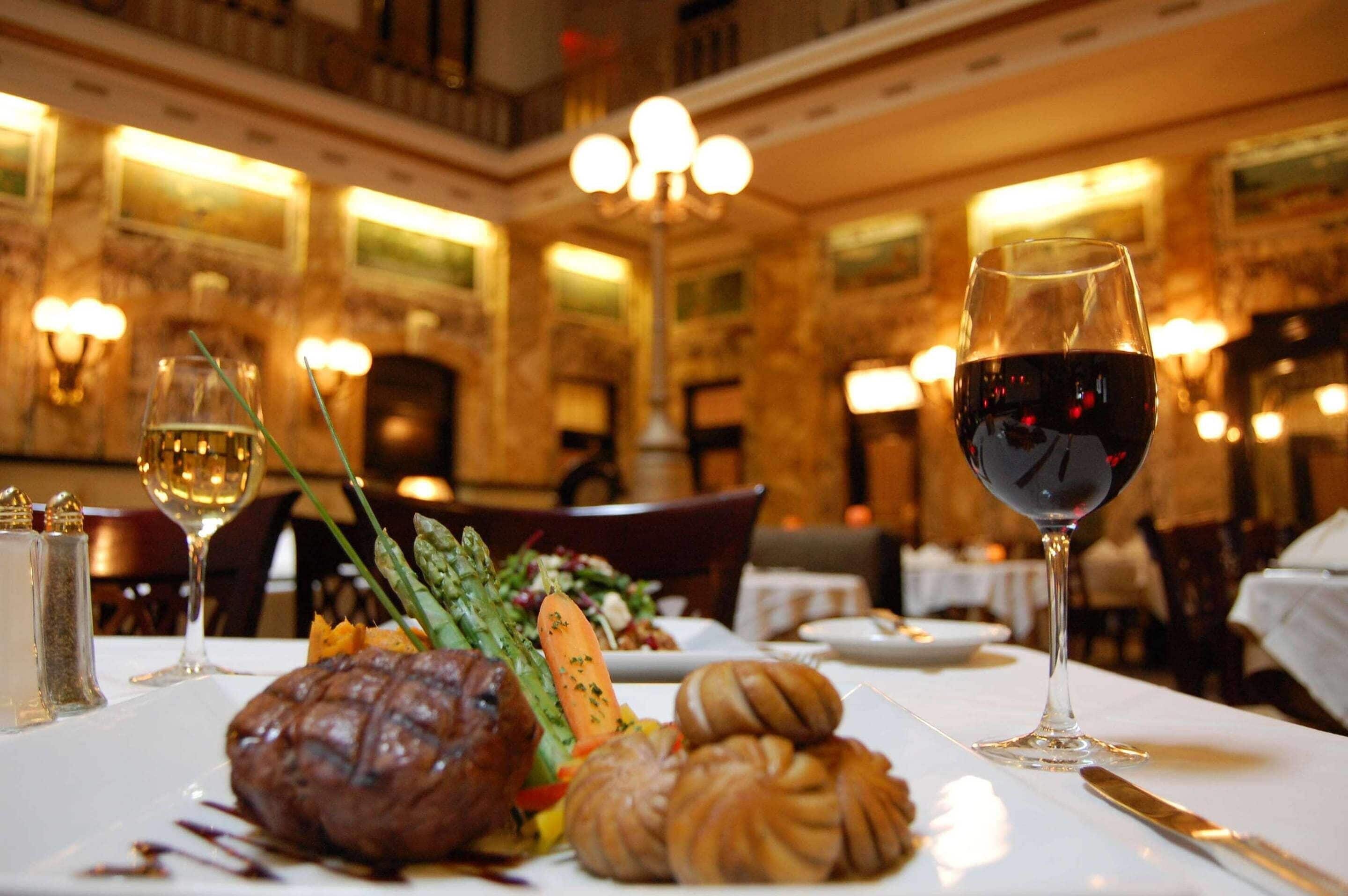 A dining table with a steak, vegetables, and pastries, and two glasses of red wine in a restaurant with ornate decor.
