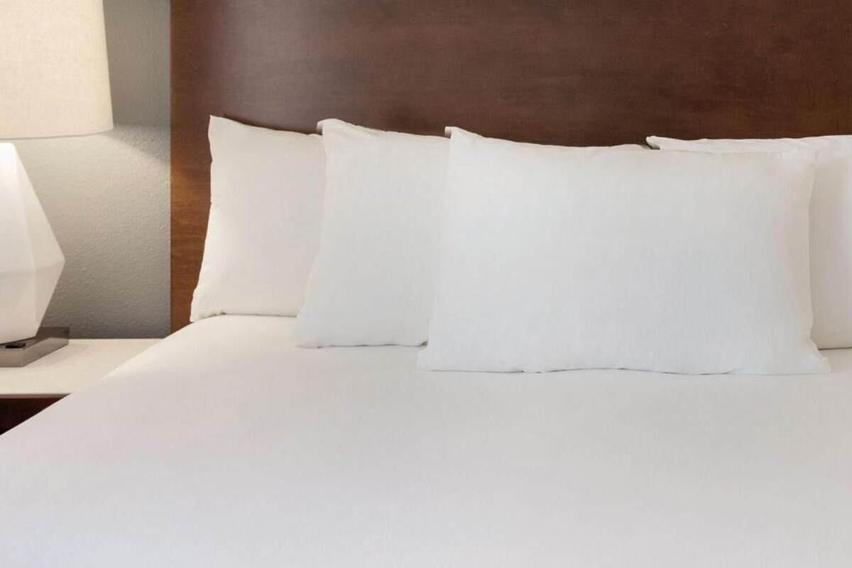 A neatly made bed with white linens and pillows, a wooden headboard, and a bedside table with a lamp.