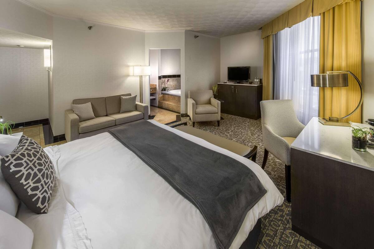 A hotel room with a large bed, a sofa, a desk, a TV, and a bathroom visible through an open door.