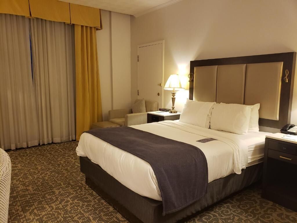 A hotel room with a large bed, a bedside table with a lamp, a chair, and a wardrobe.