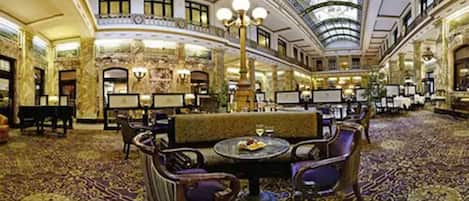 Un lobby imponente con candelabros ornamentales, una zona de estar central con una mesa dispuesta para comer, y un techo alto con tragaluz.