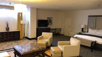 Habitación de hotel con una cama, dos sillones, una mesa de centro de madera, un televisor, un escritorio y una lámpara.