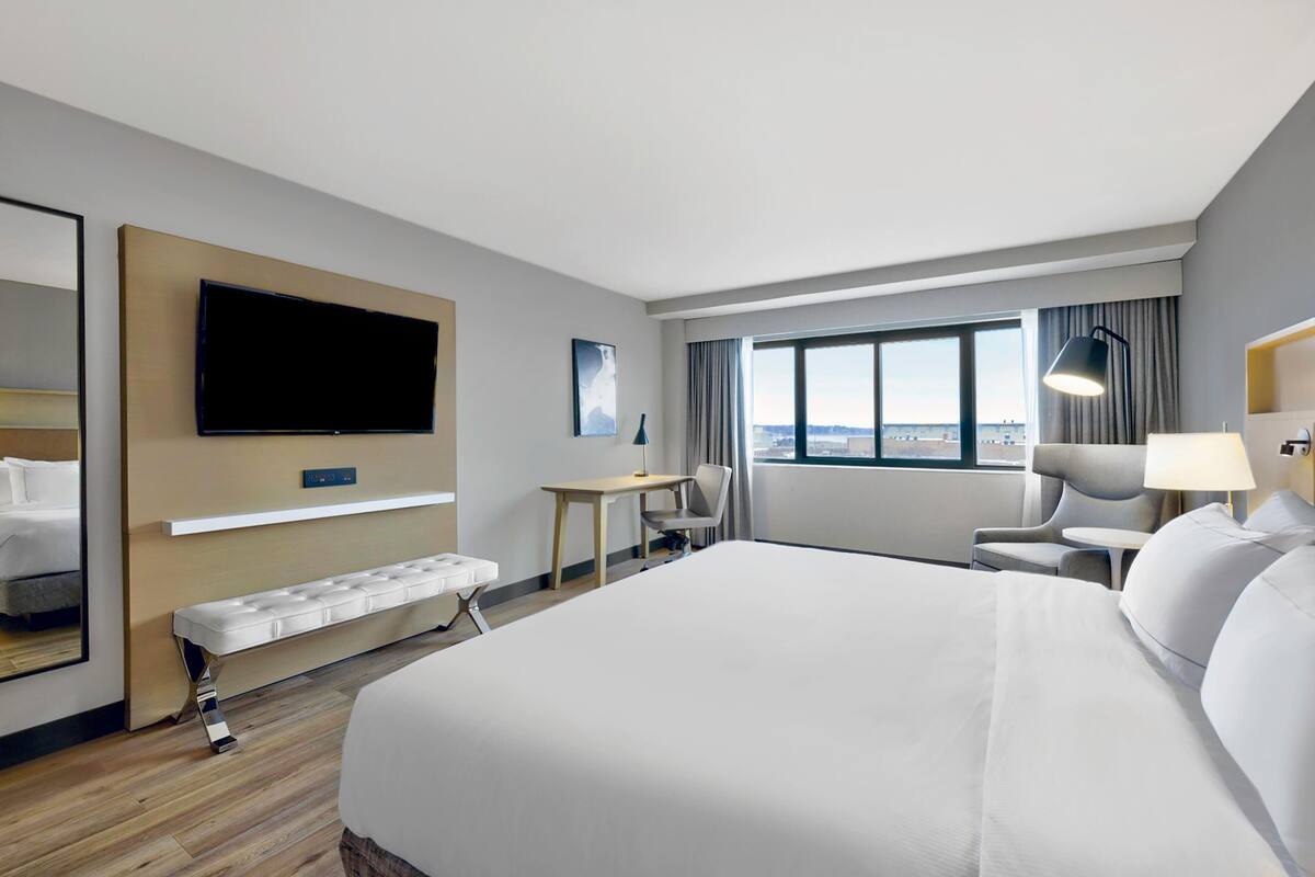 A hotel room with a large bed, a desk, a chair, a television, and a window with a city view.