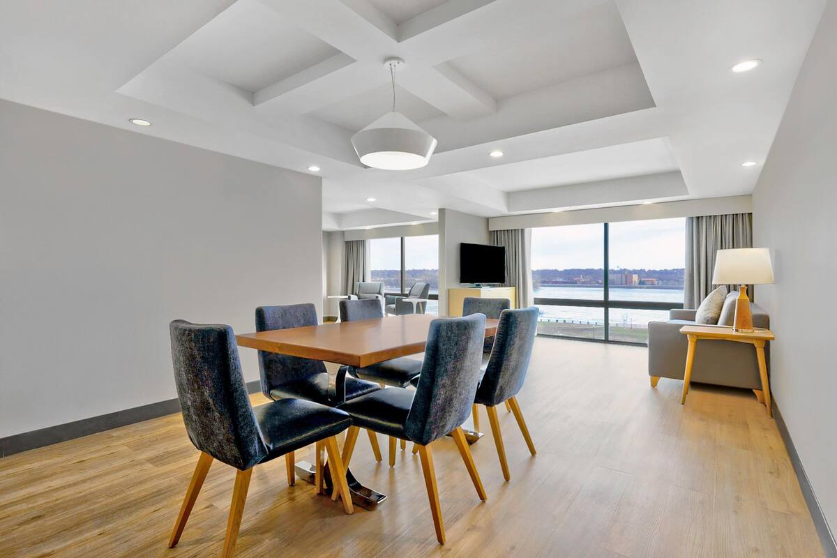 A modern hotel room with a dining area, a sofa, a TV, and a large window offering a city view.