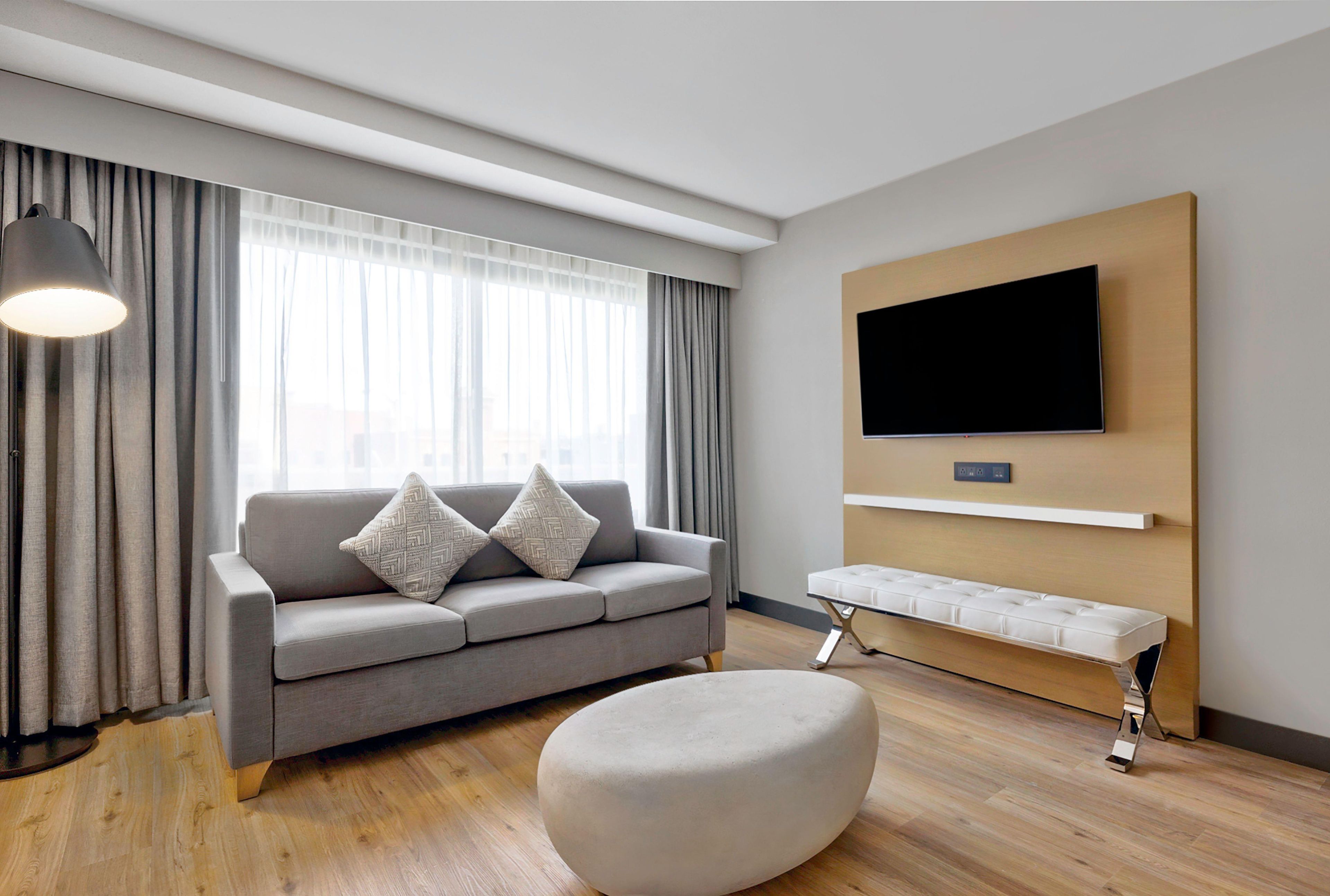 A modern living room with a grey sofa, a round ottoman, a bench, a floor lamp, and a mounted TV.