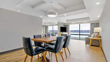 A modern hotel room with a dining area, a sofa, a TV, and a large window offering a city view.