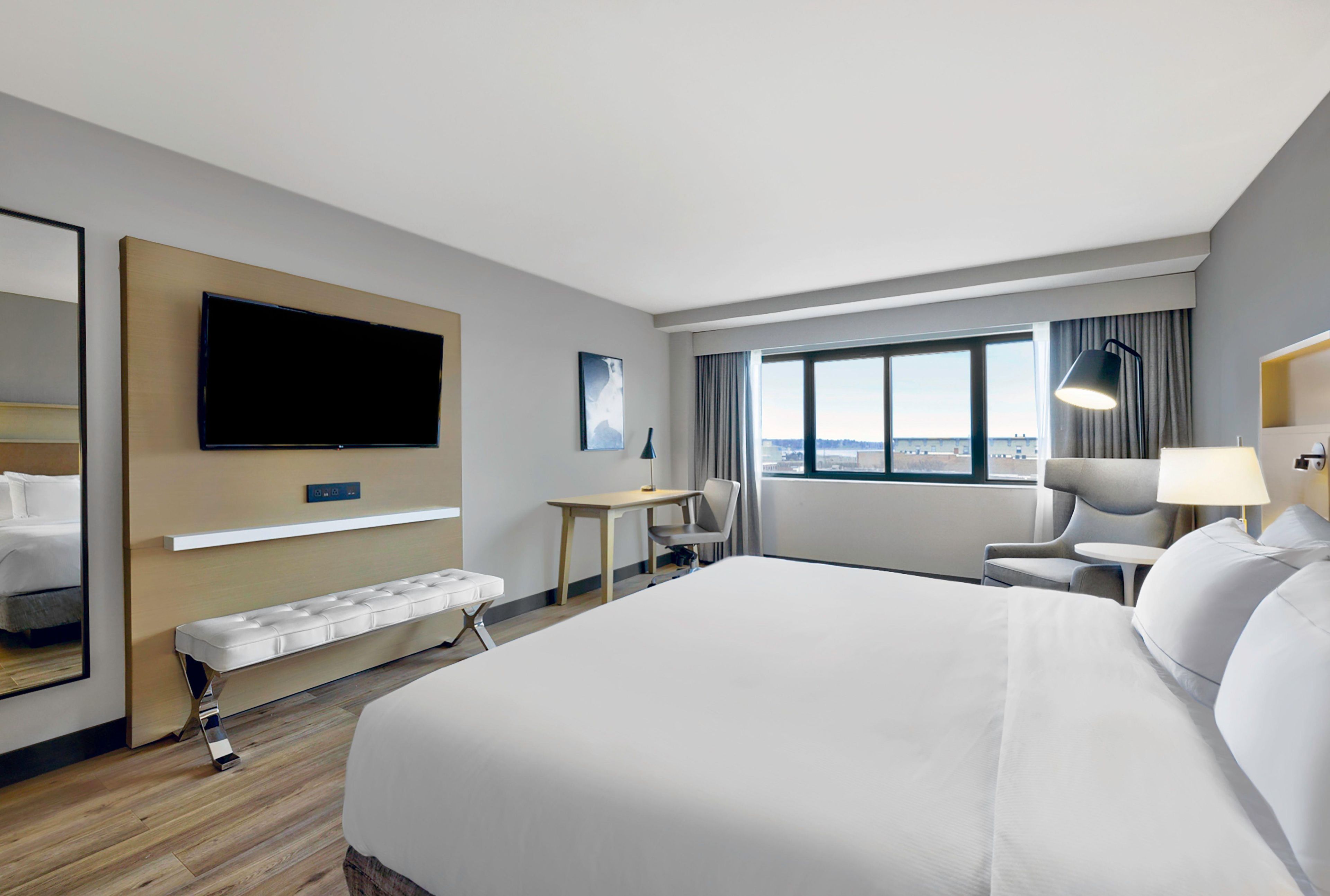 A hotel room with a large bed, a desk, a chair, a television, and a window with a city view.