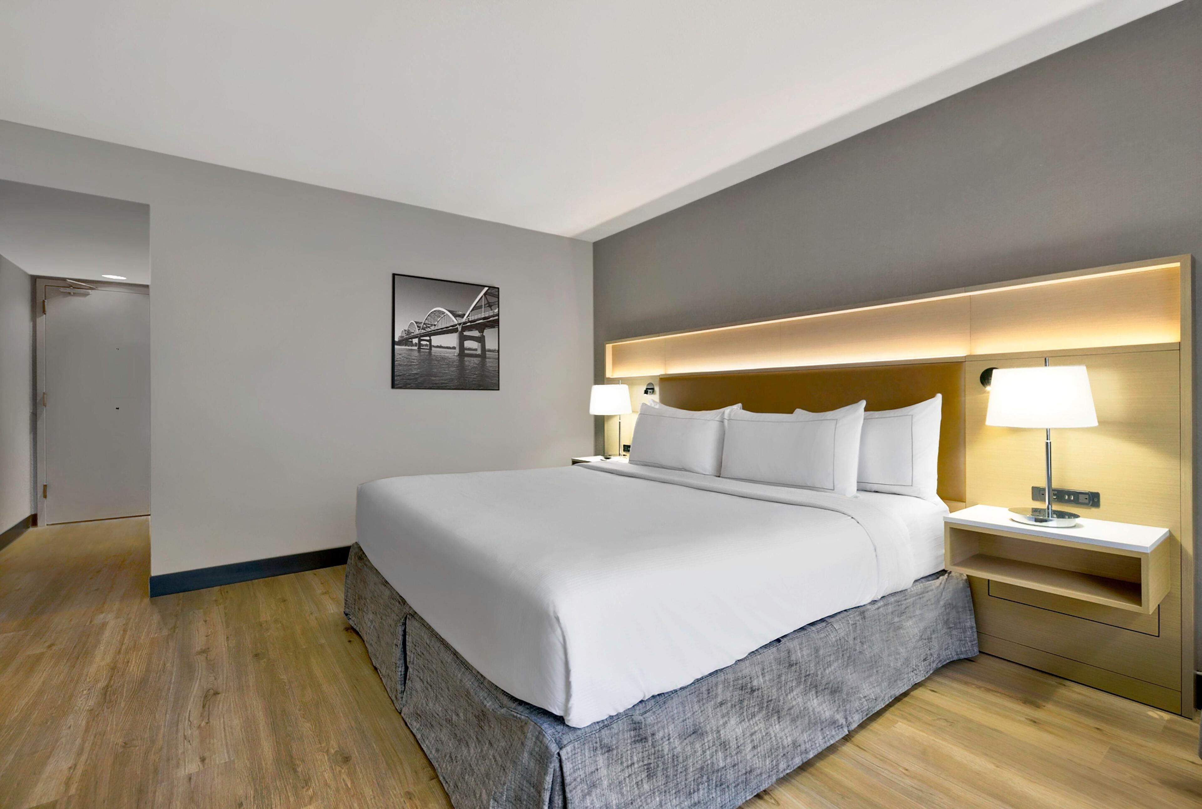 A hotel room with a large bed, a nightstand with a lamp, a framed picture on the wall, and a wooden floor.