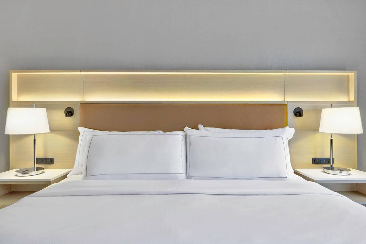 A hotel room with a large bed, two bedside tables with lamps, a headboard with integrated lighting, and a neutral-colored wall.