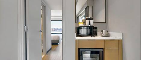 Une petite cuisine équipée d’un four à micro-ondes, d’une machine à café et d’un four, un miroir au-dessus du plan de travail et une porte donnant sur une chambre avec un lit et une fenêtre.