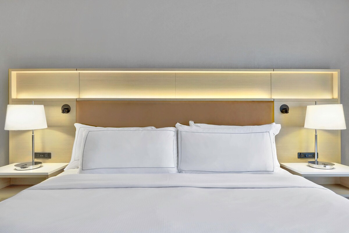 A hotel room with a large bed, two bedside tables with lamps, a headboard with integrated lighting, and a neutral-colored wall.