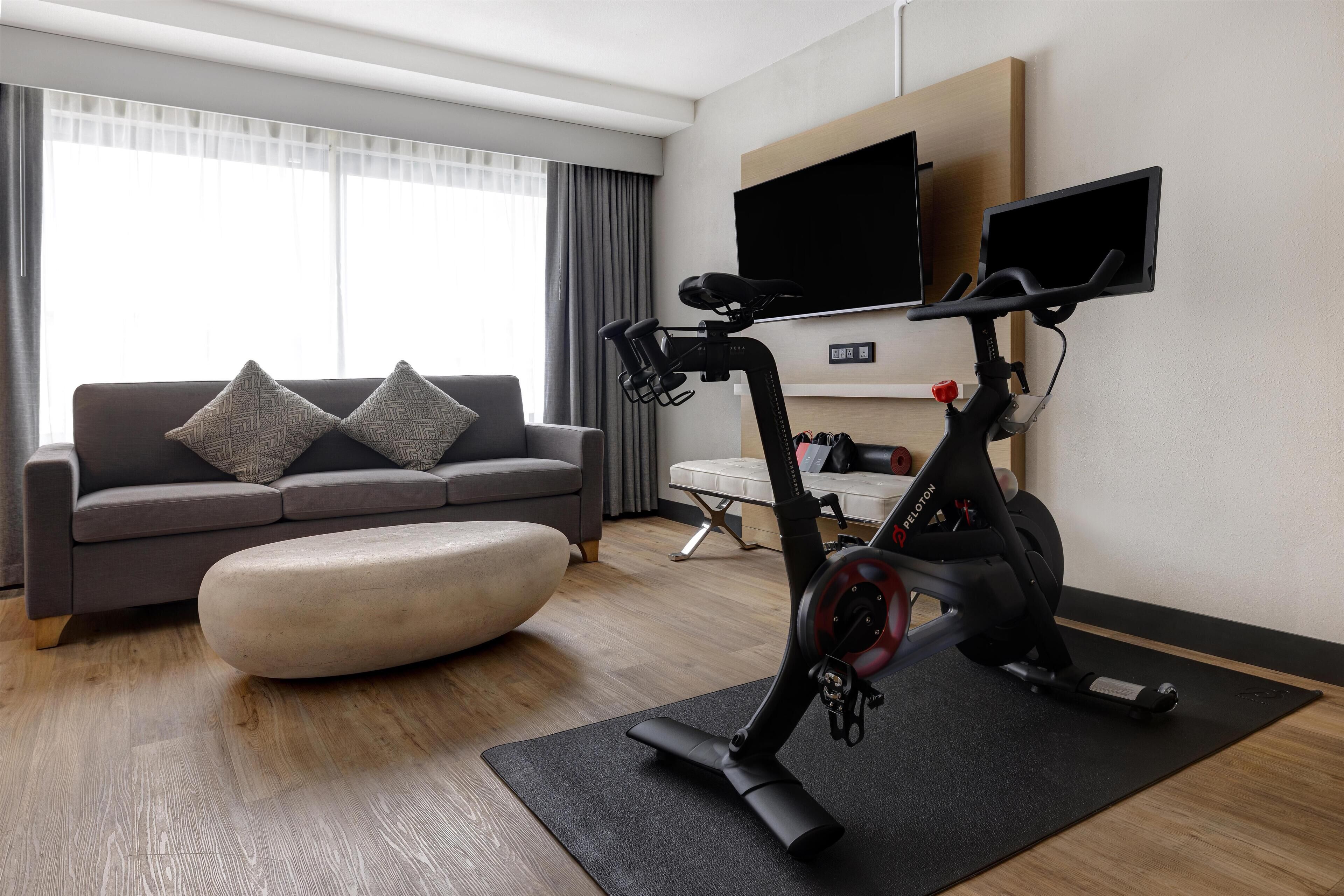 A modern living room with a grey sofa, a black exercise bike, a flat-screen TV, and a large window with curtains.