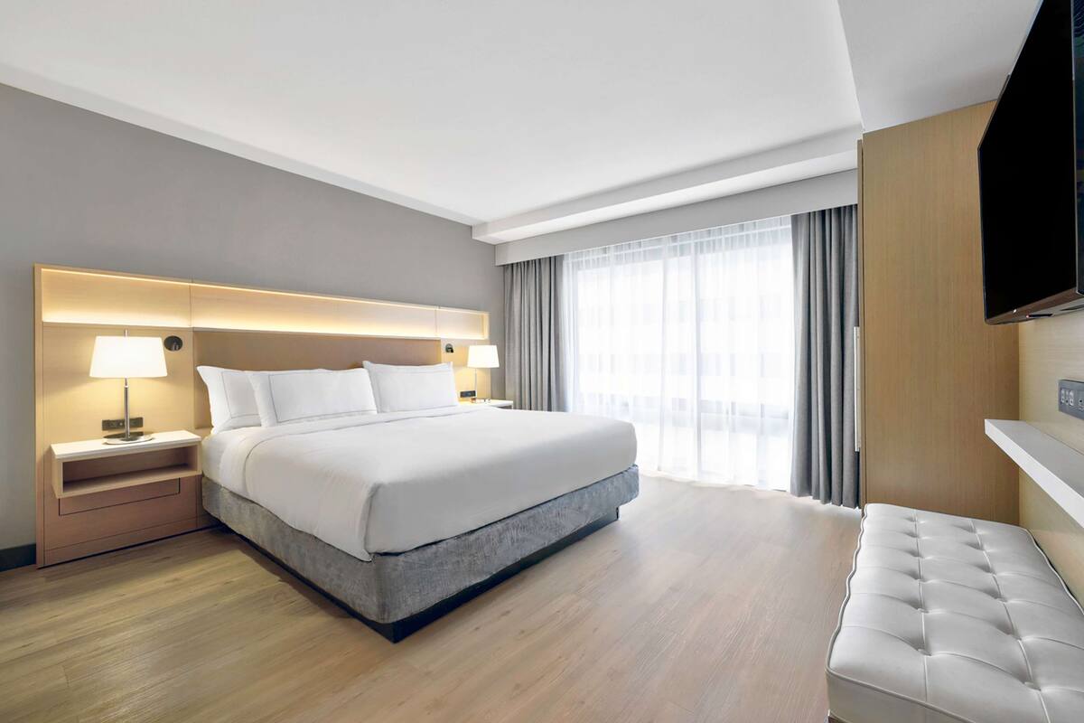A modern hotel room with a large bed, bedside lamps, a flat-screen TV, and a view of the city through large windows.