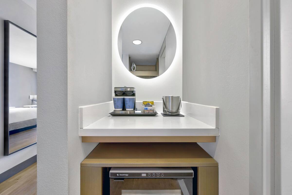 A modern hotel room with a built-in mirror, a small countertop with a coffee maker, cups, and a kettle. There is a smart fridge below the countertop.