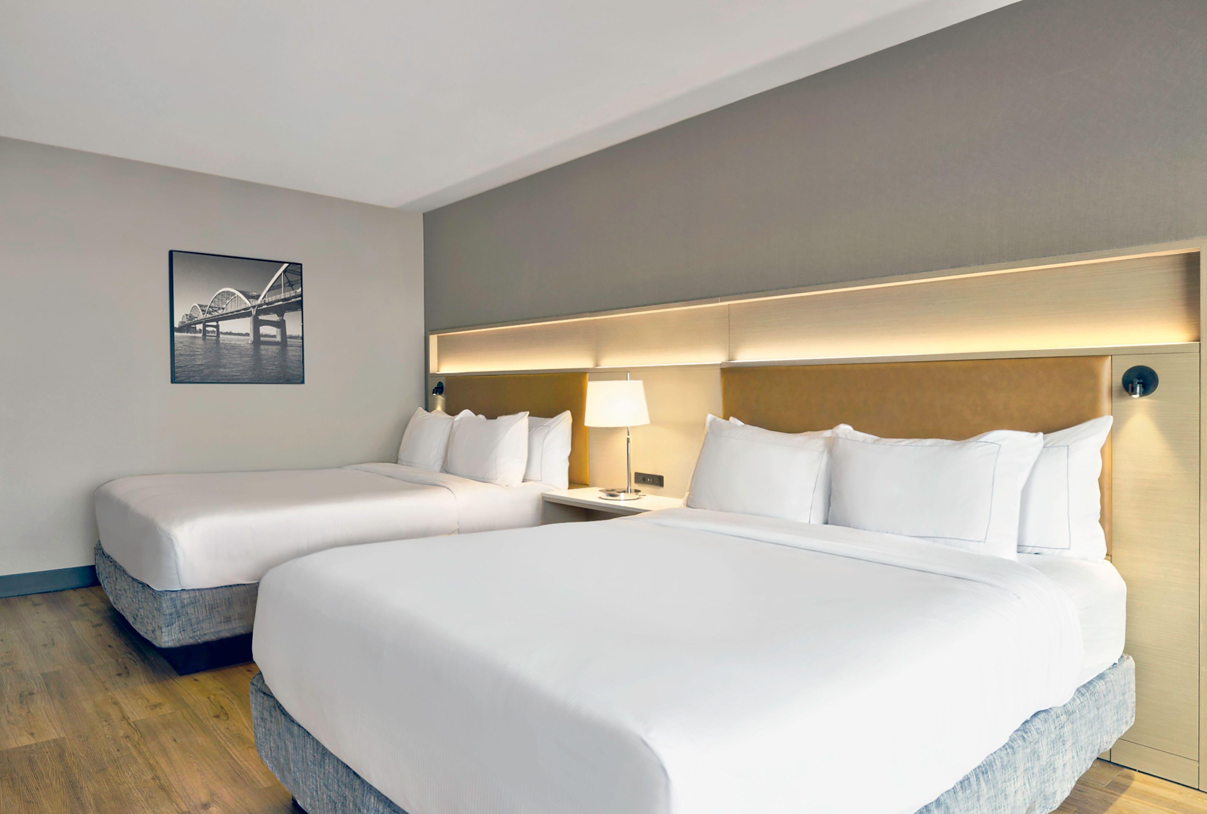 Hotellihuone, jossa on kaksi sänkyä, yöpöytä lampun kanssa ja seinällä oleva kehystetty kuva.