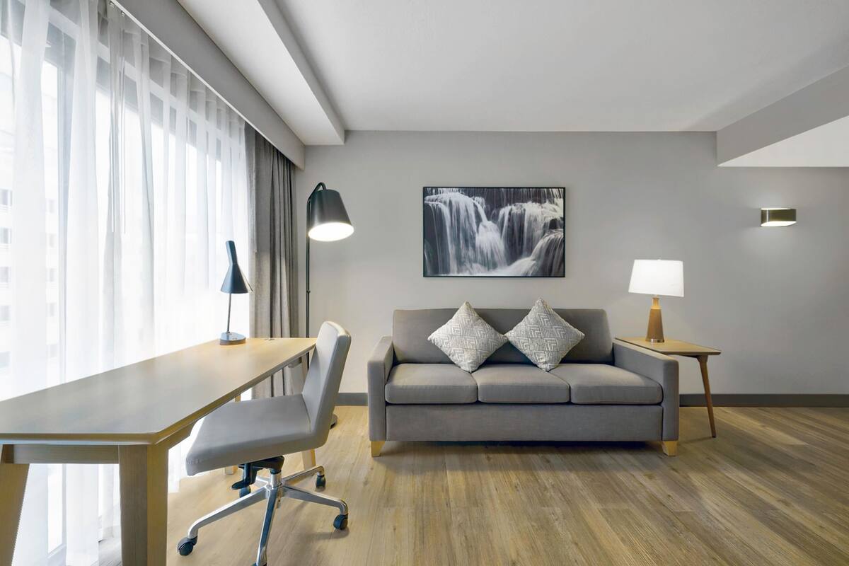 A modern hotel room with a wooden desk, a grey sofa with patterned cushions, a wall-mounted picture of a waterfall, and a small bedside table with a lamp.