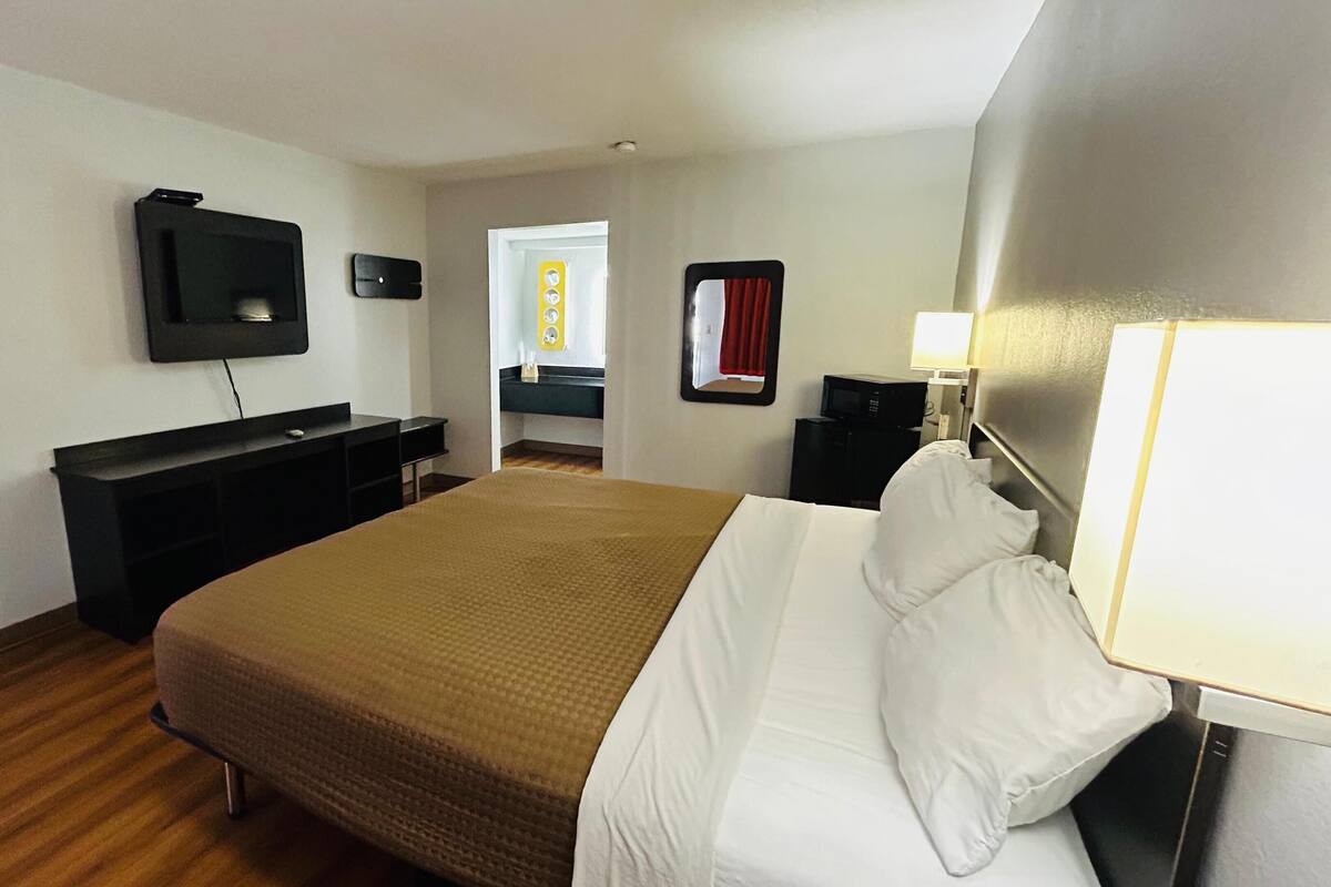 Habitación de hotel con cama, escritorio, televisor, espejo y puerta que da a un baño.