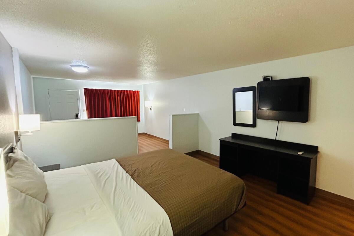 Habitación de hotel con cama, escritorio, silla, televisor y ventana con cortinas rojas.