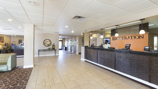 A hotel reception area with a registration desk, a seating area with a patterned rug, and a hallway leading to elevators. - Rosen Inn, closest to Universal (Orlando)