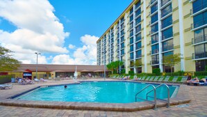 A hotel pool area with a rectangular pool, lounge chairs, and a multi-story building with balconies.
