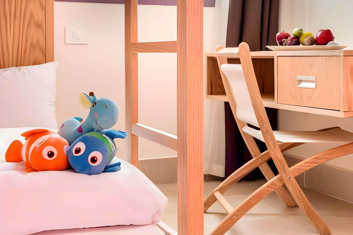 A bedroom with a bed, a desk with a fruit bowl, and plush toys.