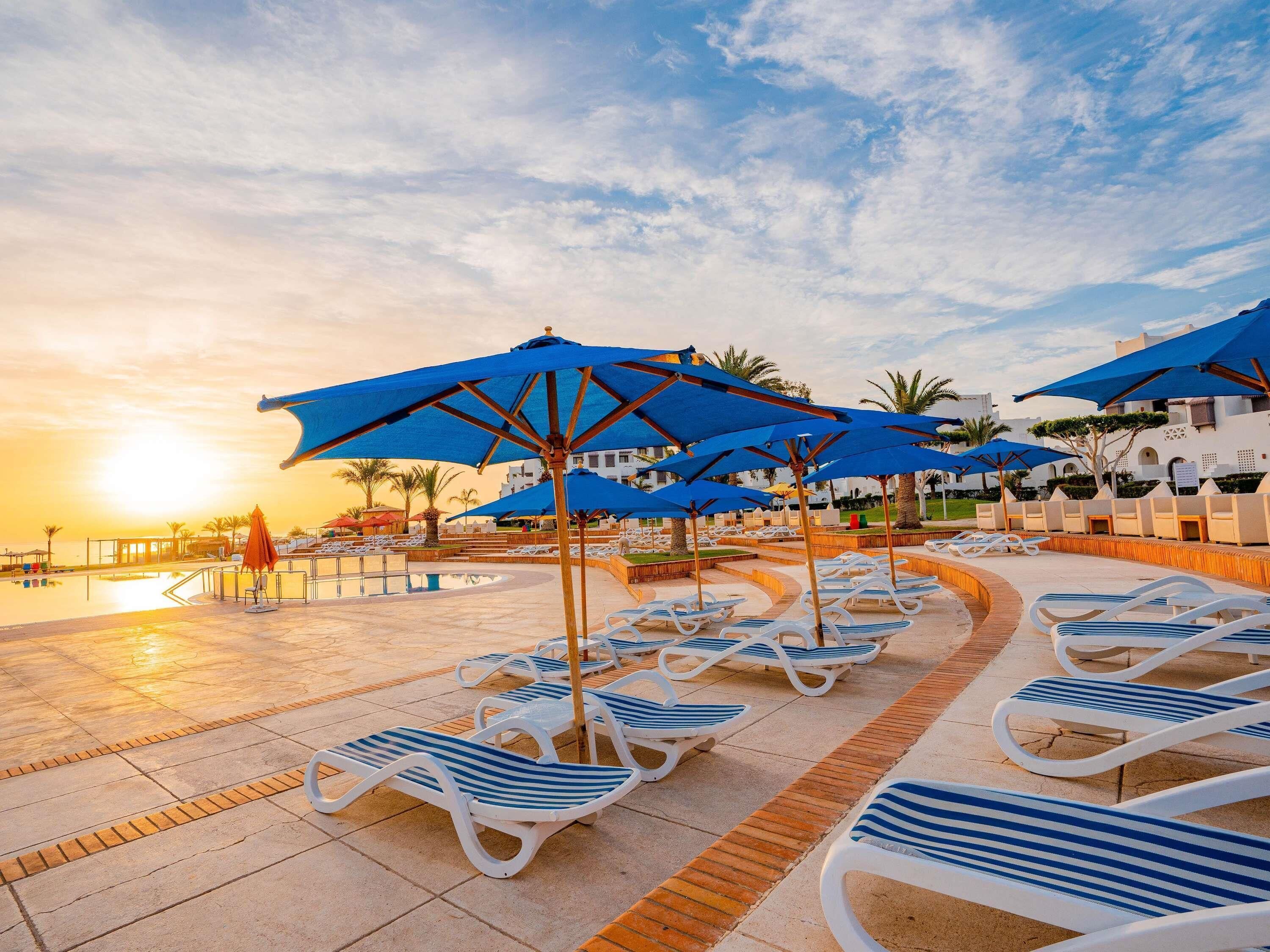 A poolside area with lounge chairs and blue umbrellas, a clear sky, and a sunset.