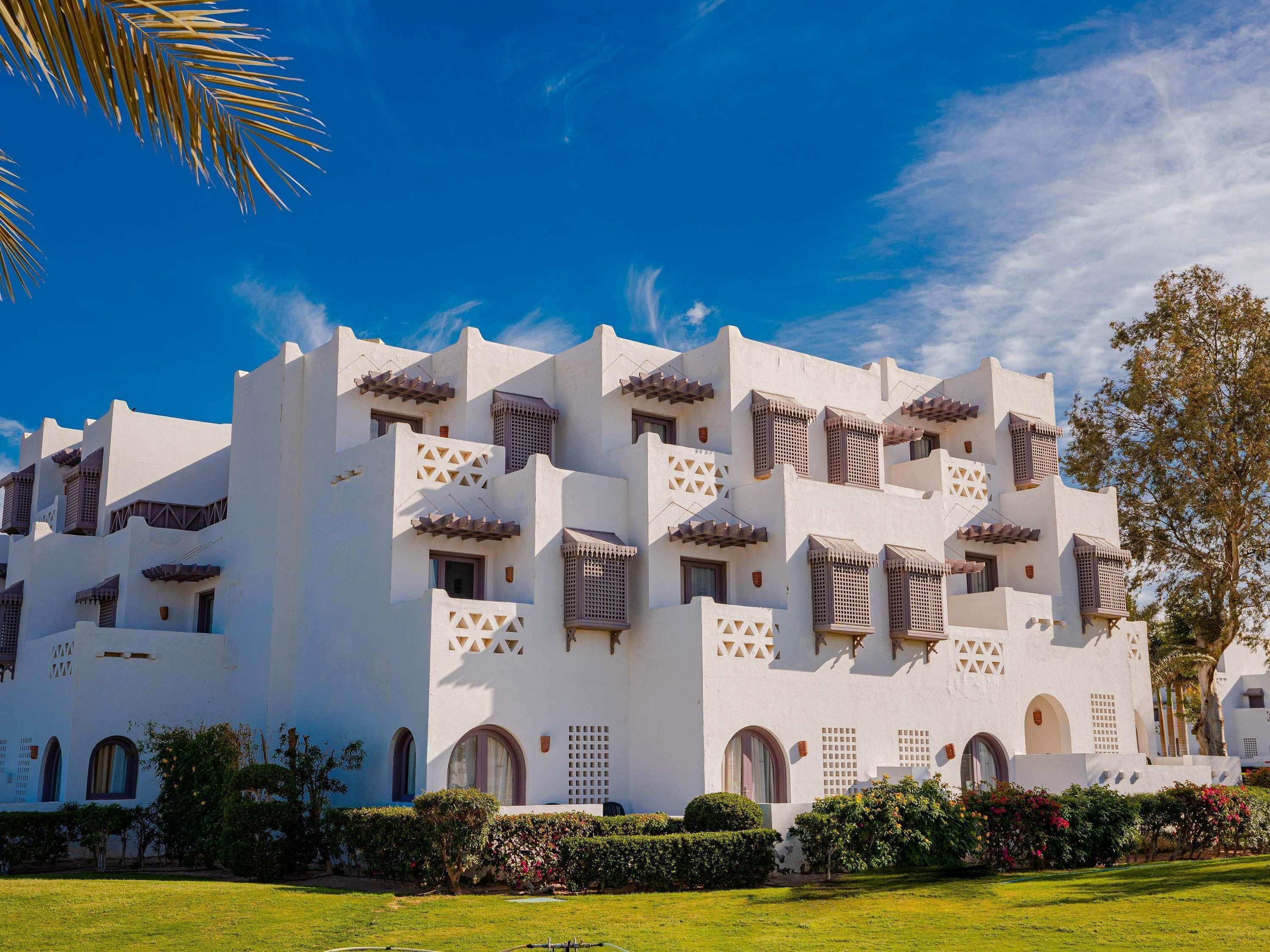 A white building with multiple balconies and arched windows, surrounded by well-maintained greenery.