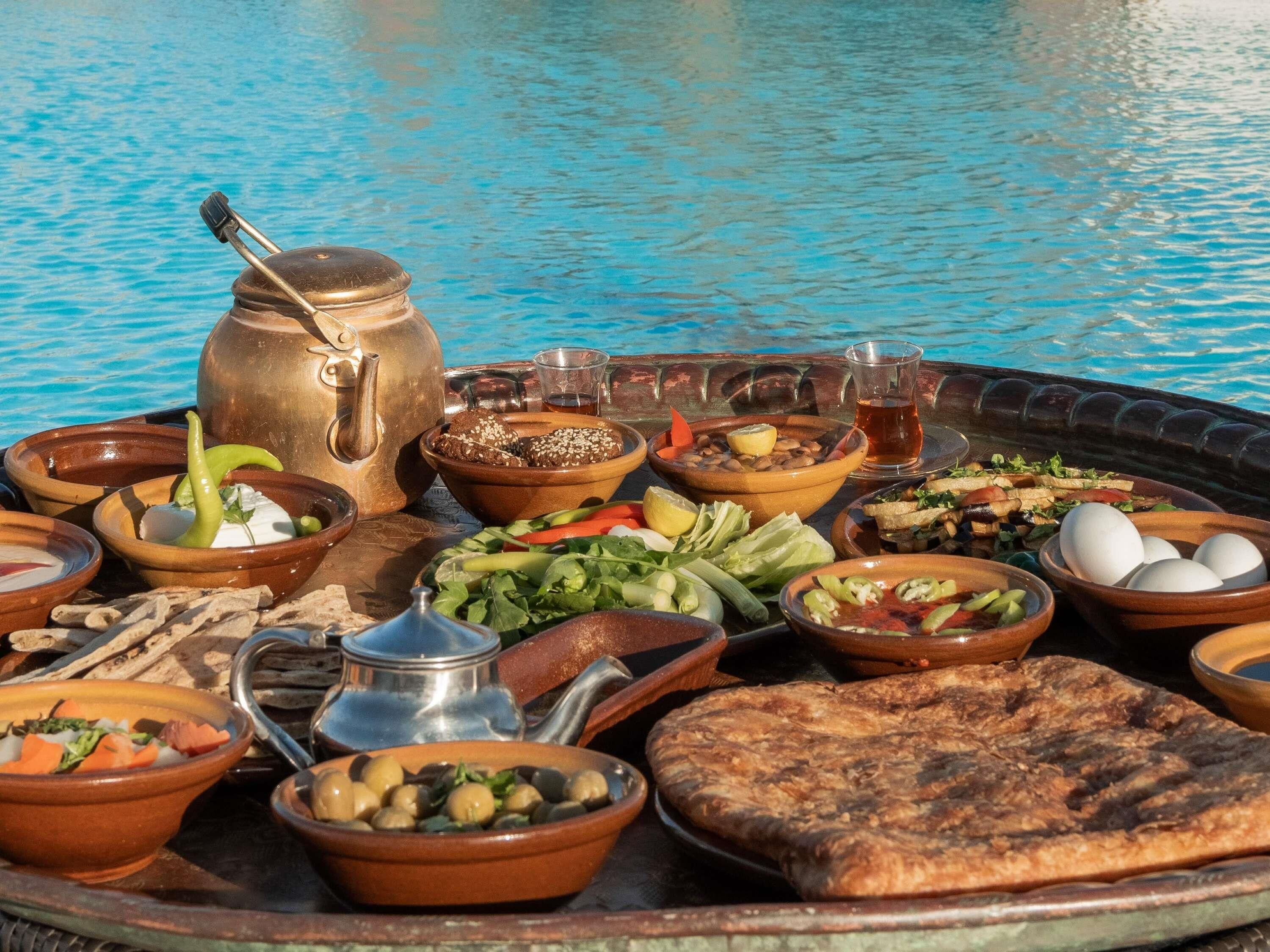Een tafel met diverse gerechten en een theepot bij een zwembad.